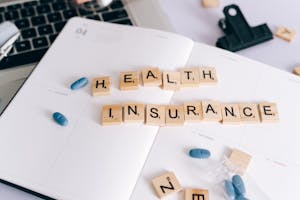 Scrabble tiles spelling 'Health Insurance' on planner with pills and laptop, symbolizing healthcare planning.